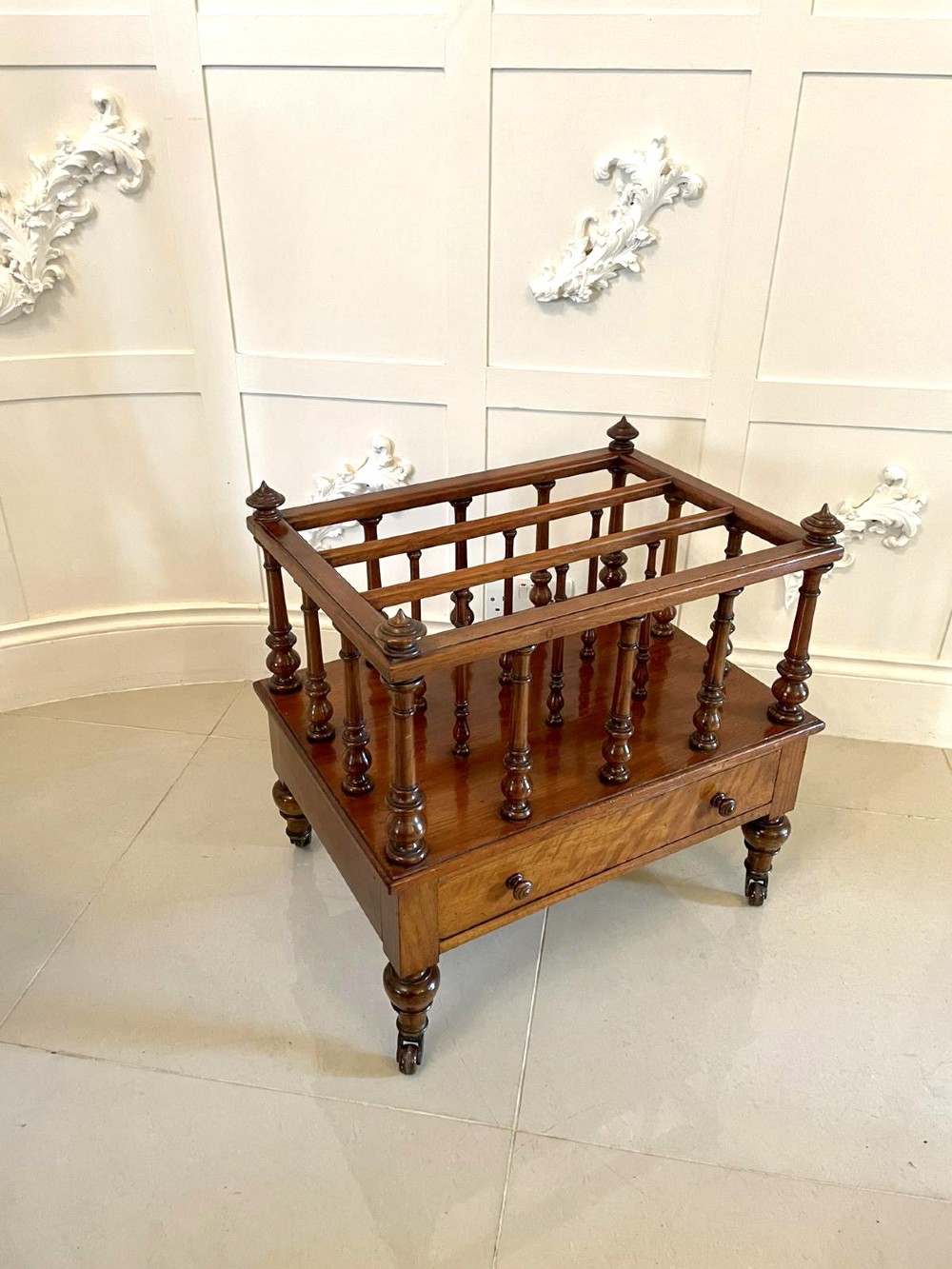 A Victorian figured walnut Canterbury featuring multiple vertical turned spindles, corner finials, a lower base drawer with two knobs, and brass castors. View XIII