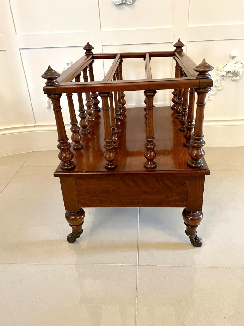 A Victorian figured walnut Canterbury featuring multiple vertical turned spindles, corner finials, a lower base drawer with two knobs, and brass castors. View XIV