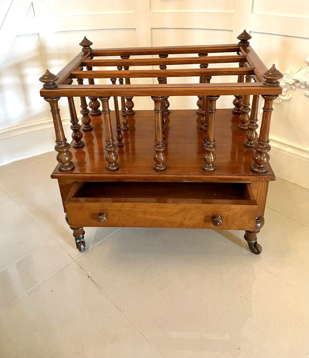 A Victorian figured walnut Canterbury featuring multiple vertical turned spindles, corner finials, a lower base drawer with two knobs, and brass castors. View XV