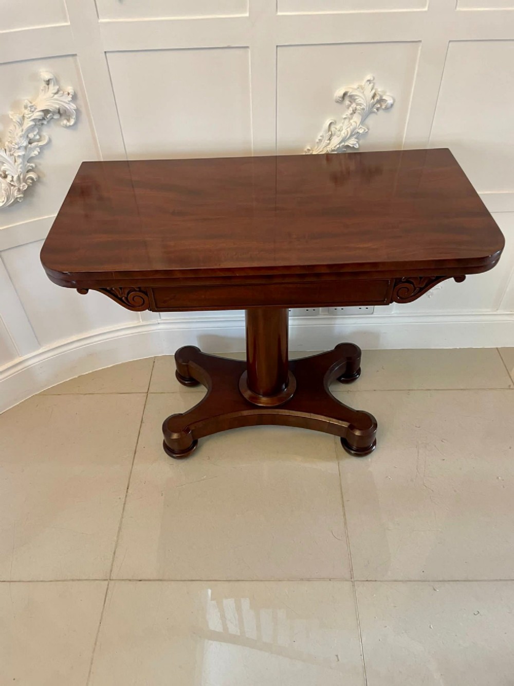 A Victorian mahogany card table featuring a rectangular fold-over top, carved scroll apron brackets, and a central cylindrical pedestal on a four-point plinth base. View IX