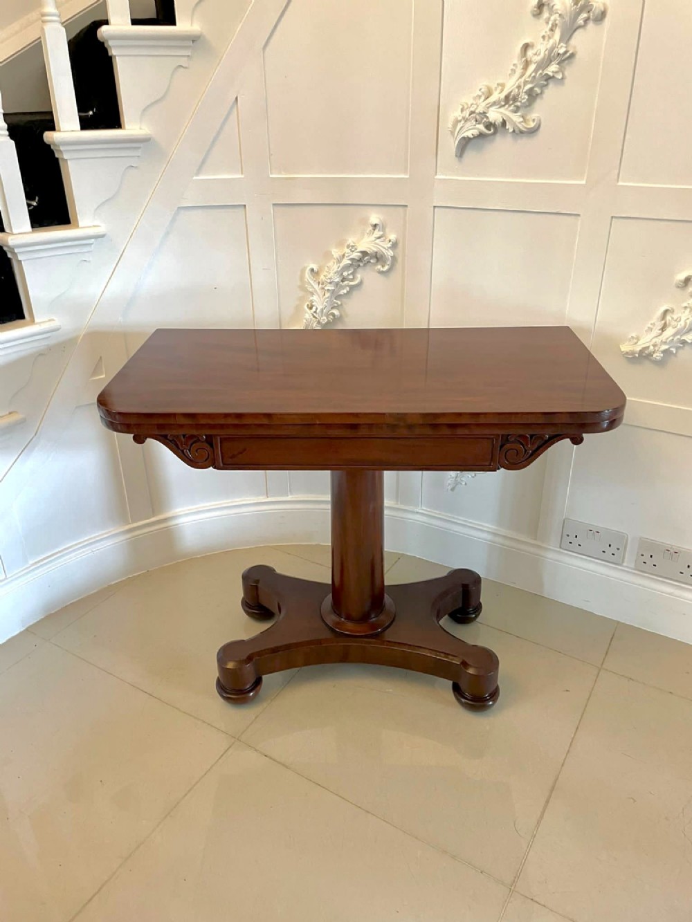 A Victorian mahogany card table featuring a rectangular fold-over top, carved scroll apron brackets, and a central cylindrical pedestal on a four-point plinth base. View X