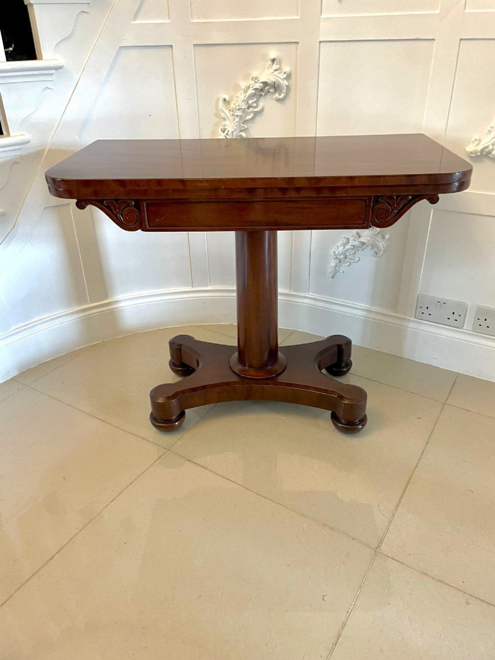 A Victorian mahogany card table featuring a rectangular fold-over top, carved scroll apron brackets, and a central cylindrical pedestal on a four-point plinth base. View XII
