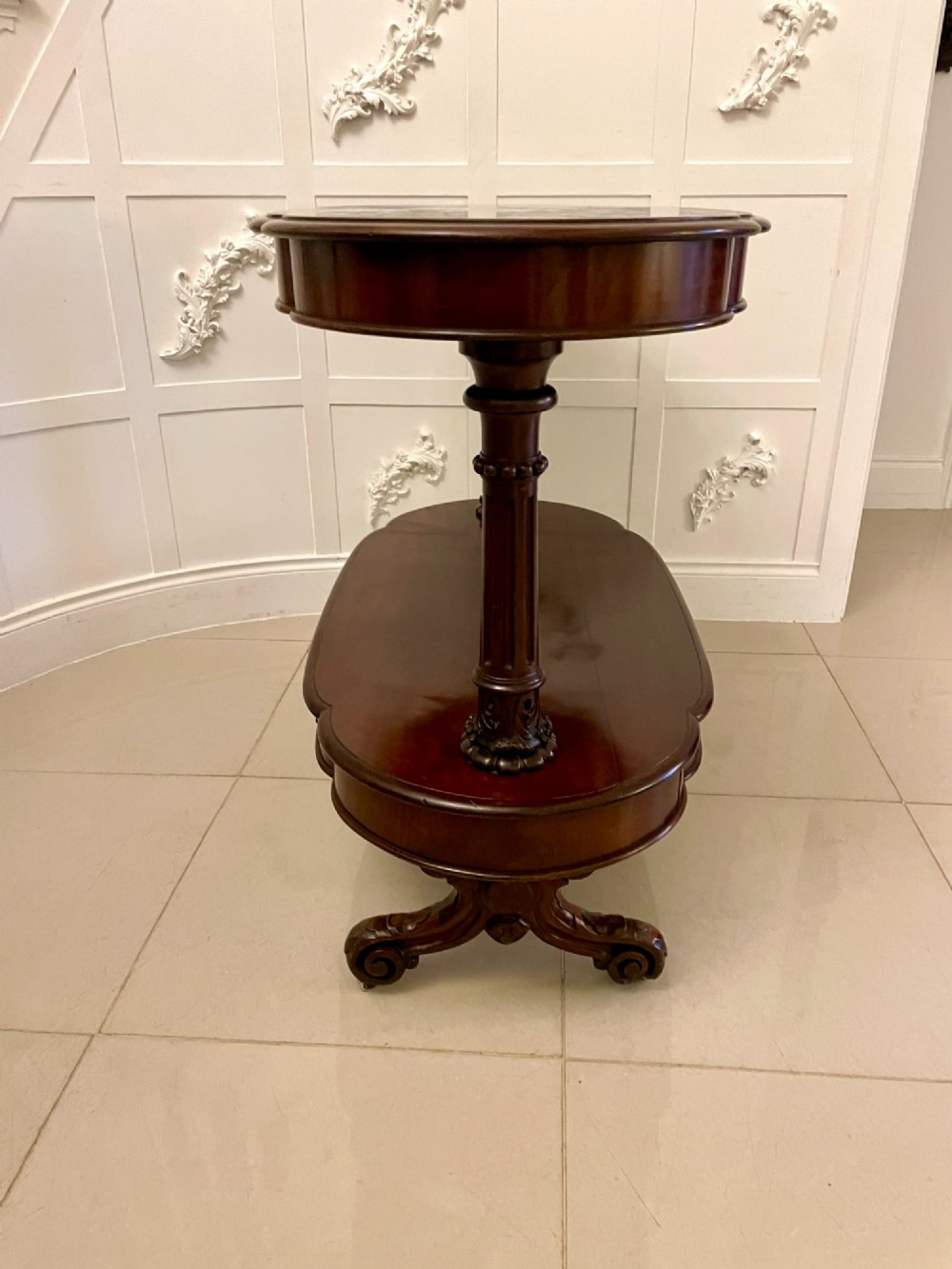 A two-tiered oval mahogany console table featuring carved column supports, a smooth polished finish, and castors on scroll feet. View III