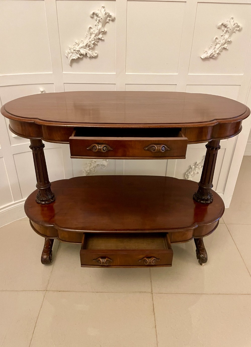 A two-tiered oval mahogany console table featuring carved column supports, a smooth polished finish, and castors on scroll feet. View V