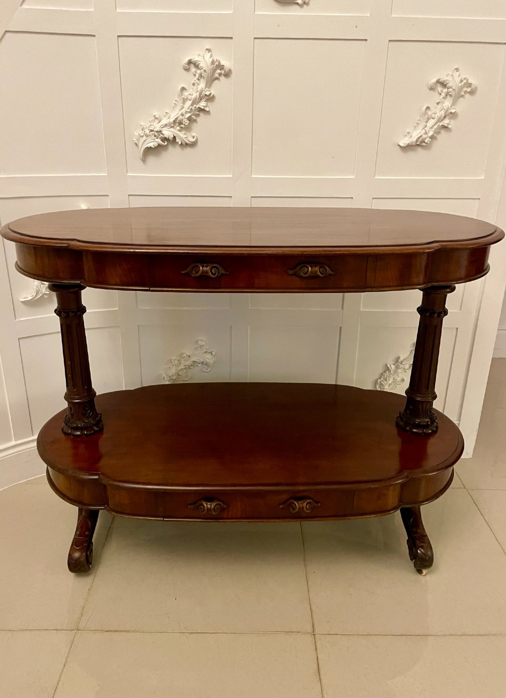 A two-tiered oval mahogany console table featuring carved column supports, a smooth polished finish, and castors on scroll feet. View XIII