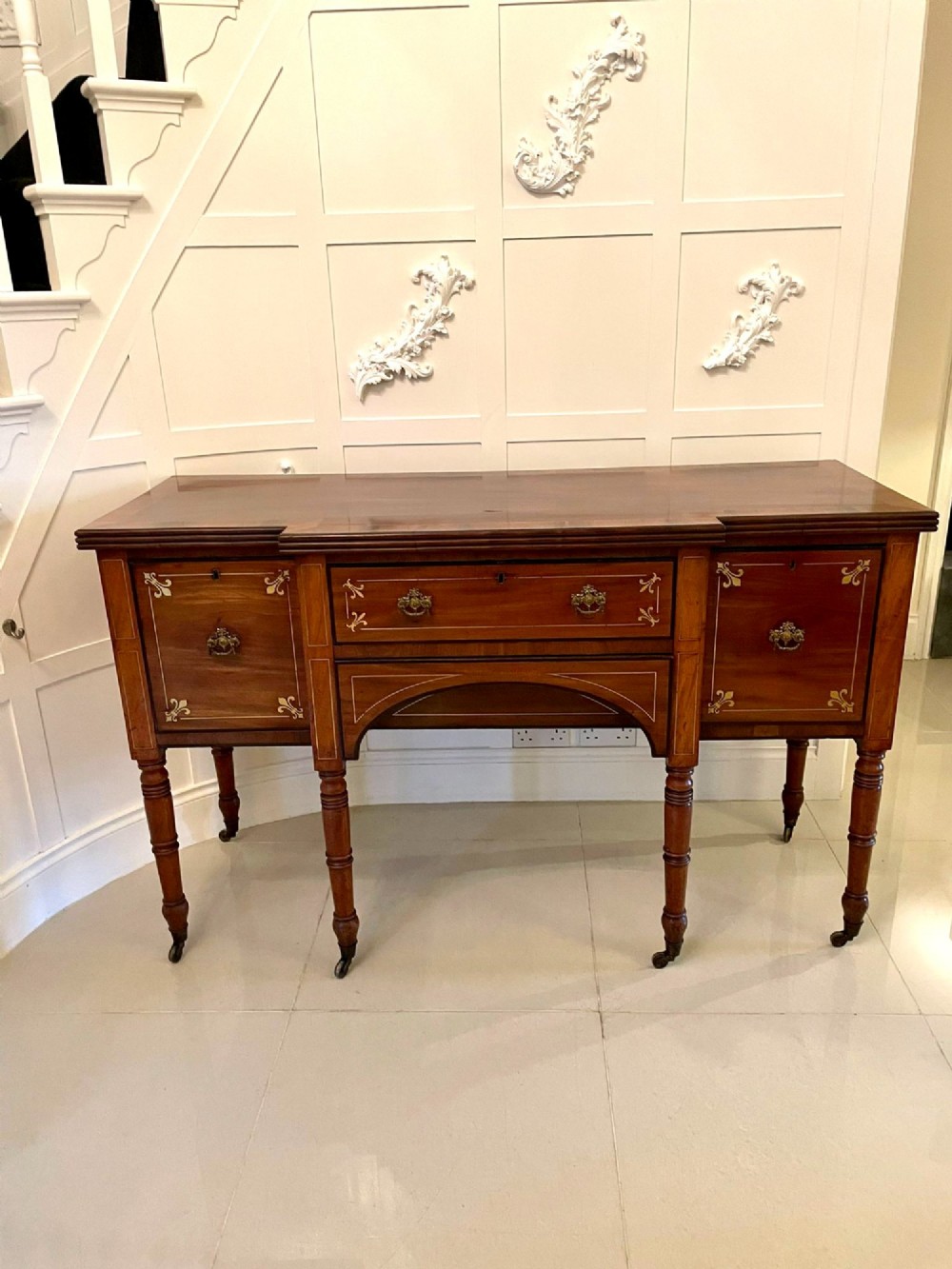 A Regency period mahogany breakfront sideboard featuring brass inlay motifs, a central drawer flanked by cupboards, turned legs, and brass castors.