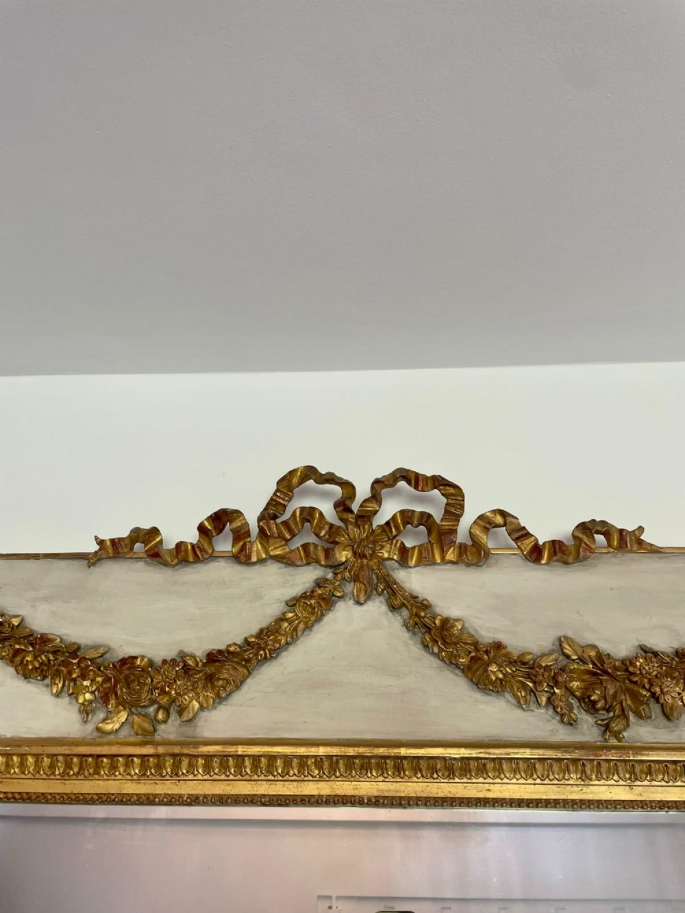 A tall rectangular Victorian mirror featuring a white painted outer frame, gilded beaded inner border, and a gilded ribbon crest with floral swags. View V