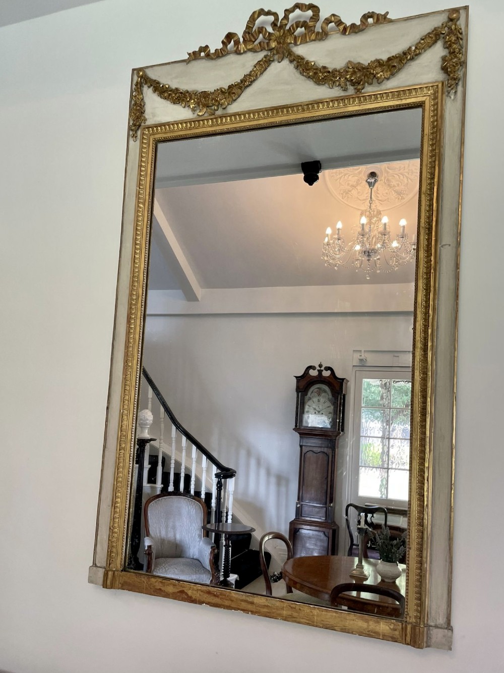 A tall rectangular Victorian mirror featuring a white painted outer frame, gilded beaded inner border, and a gilded ribbon crest with floral swags. View VI