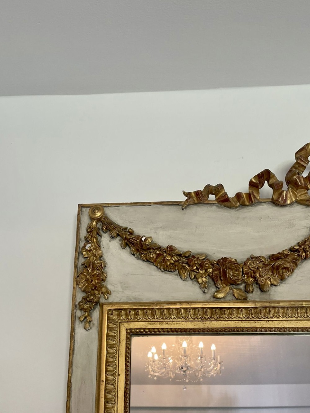 A tall rectangular Victorian mirror featuring a white painted outer frame, gilded beaded inner border, and a gilded ribbon crest with floral swags. View VII