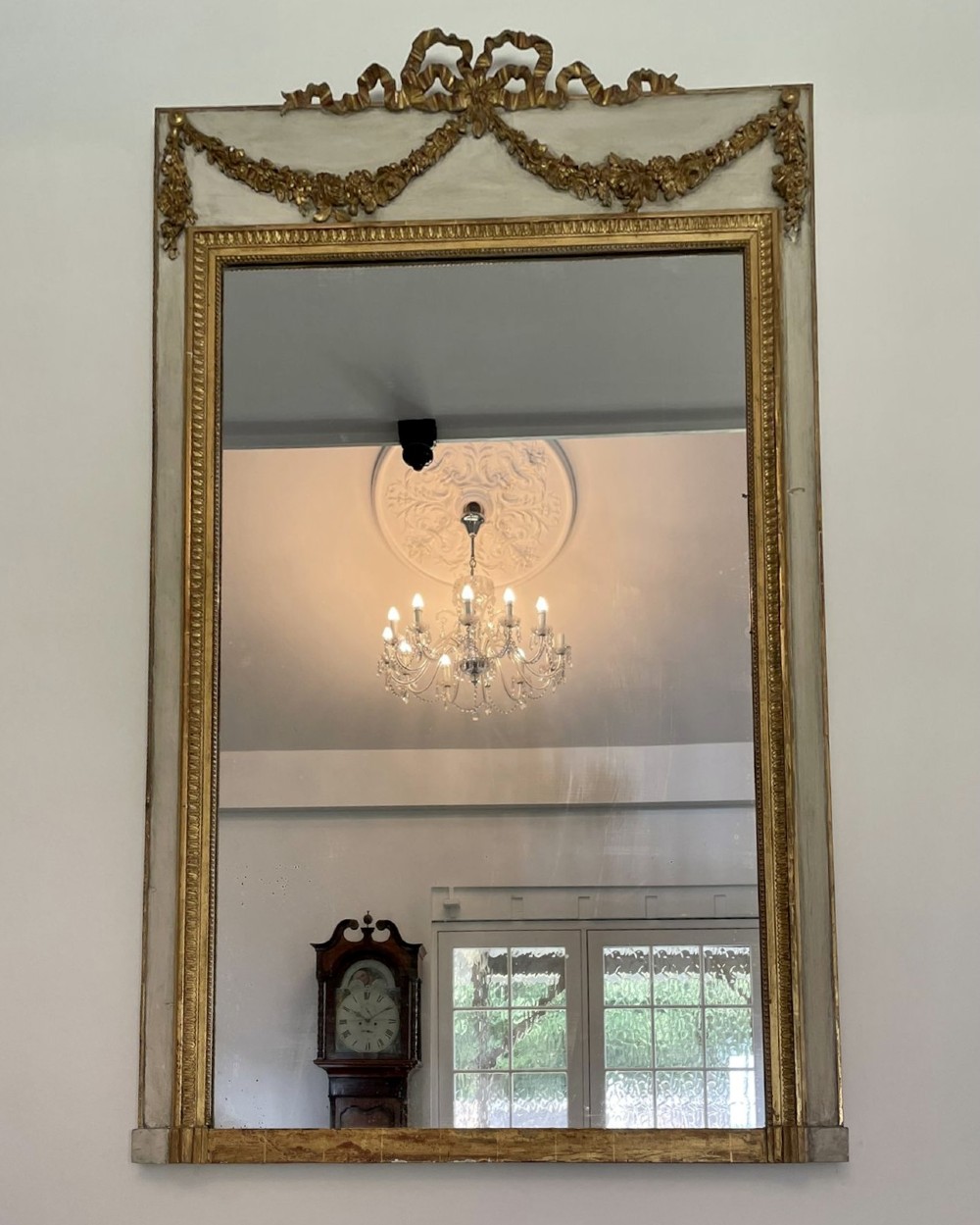 A tall rectangular Victorian mirror featuring a white painted outer frame, gilded beaded inner border, and a gilded ribbon crest with floral swags.