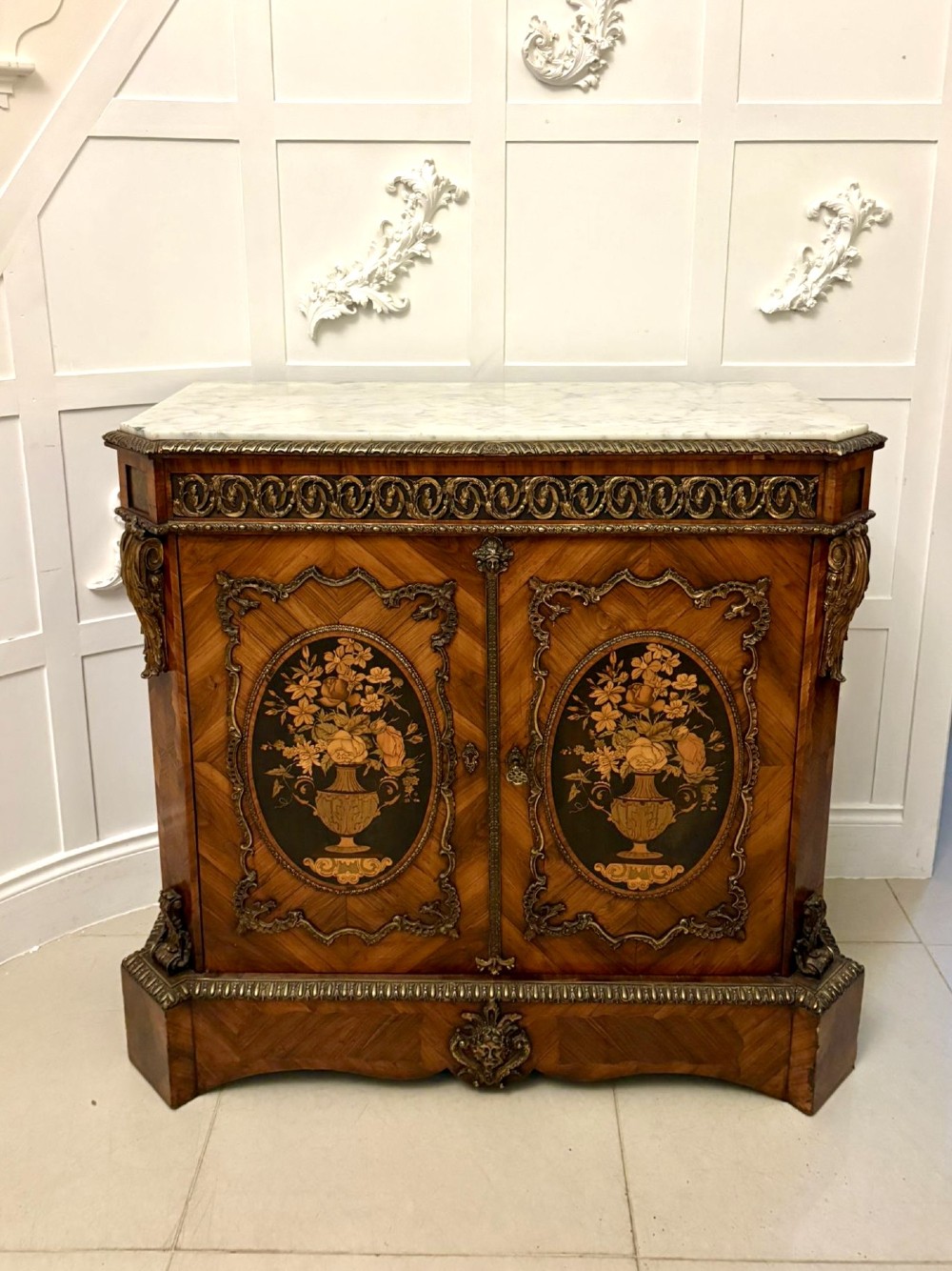 A French Louis XVI style wooden side cabinet featuring a white marble top, gilt-bronze mounts, and two door panels decorated with floral marquetry.