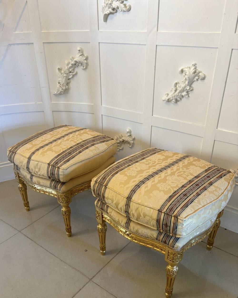 A pair of square French style footstools featuring carved gold giltwood frames, fluted legs, and yellow patterned upholstery.