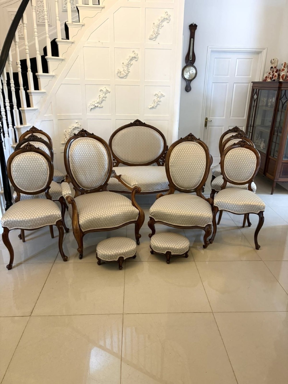 An eleven-piece antique Victorian carved walnut salon suite featuring a central sofa, pair of armchairs, six side chairs, and two footstools, all upholstered in a patterned cream fabric.