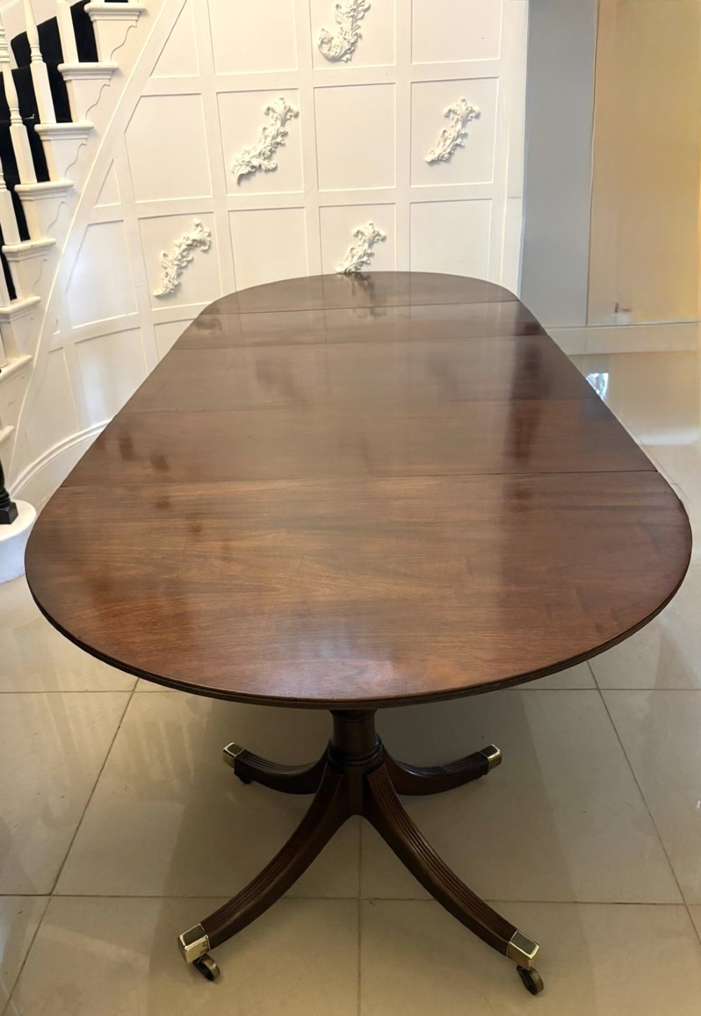 A large mahogany extending dining table featuring an oval surface with two additional leaves, supported by splayed pedestal legs with brass castors. View VI