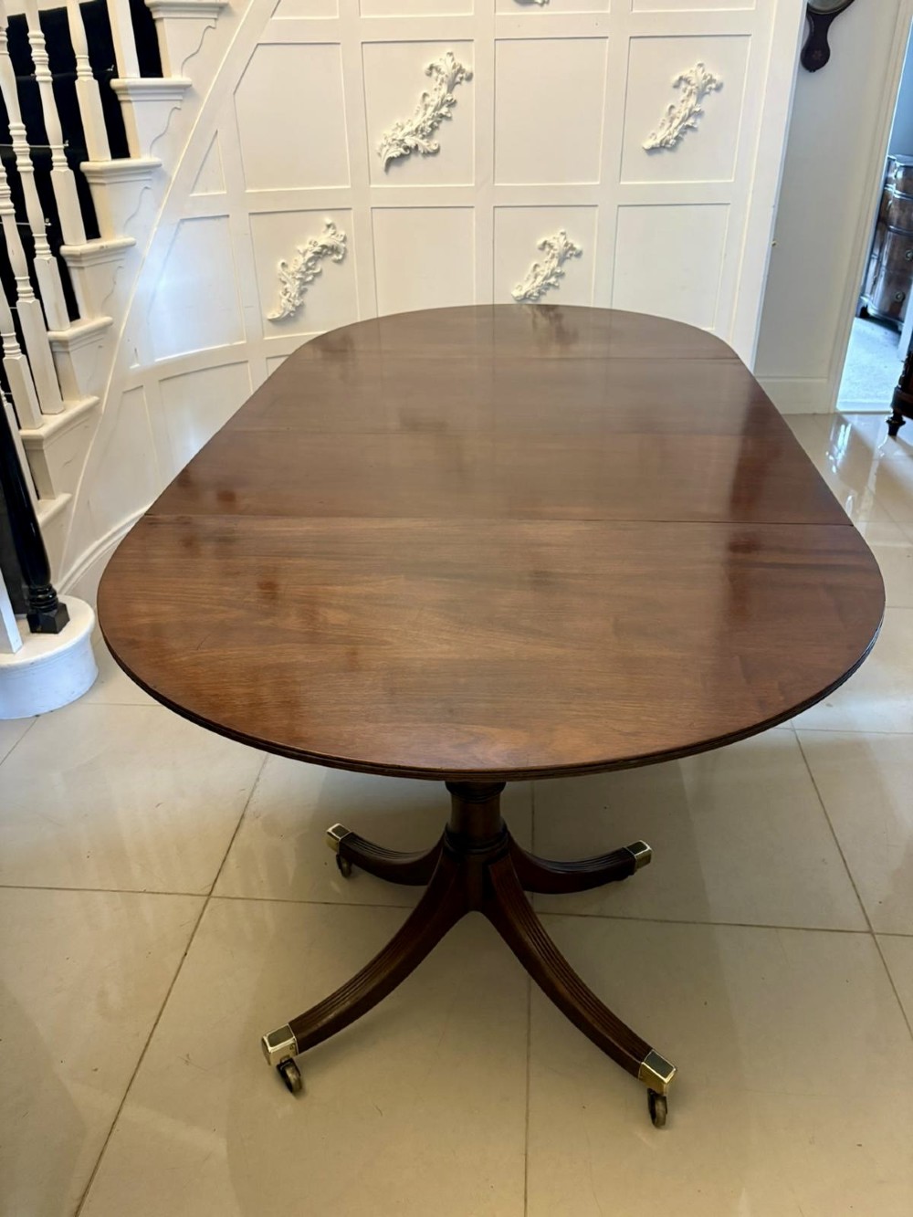 A large mahogany extending dining table featuring an oval surface with two additional leaves, supported by splayed pedestal legs with brass castors. View IX