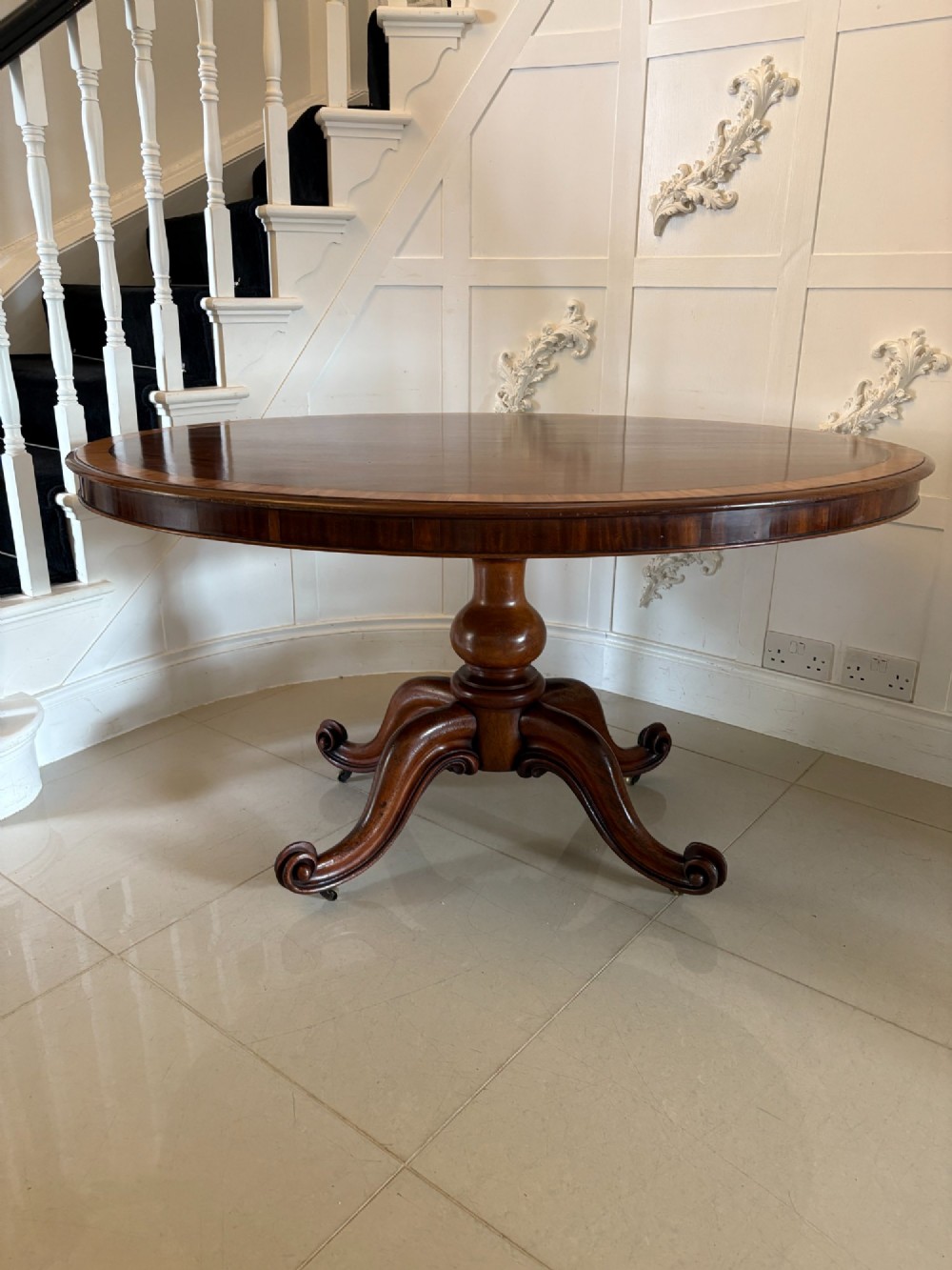 An oval Victorian tilt-top dining table in figured mahogany with satinwood crossbanding, set on a quadruped pedestal base with brass castors. View II