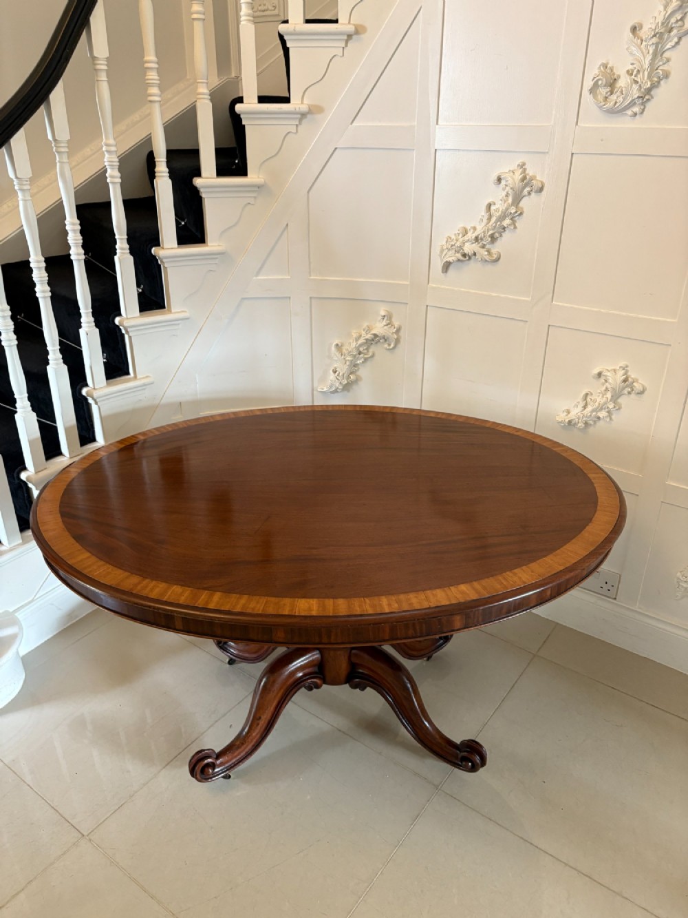 An oval Victorian tilt-top dining table in figured mahogany with satinwood crossbanding, set on a quadruped pedestal base with brass castors. View III