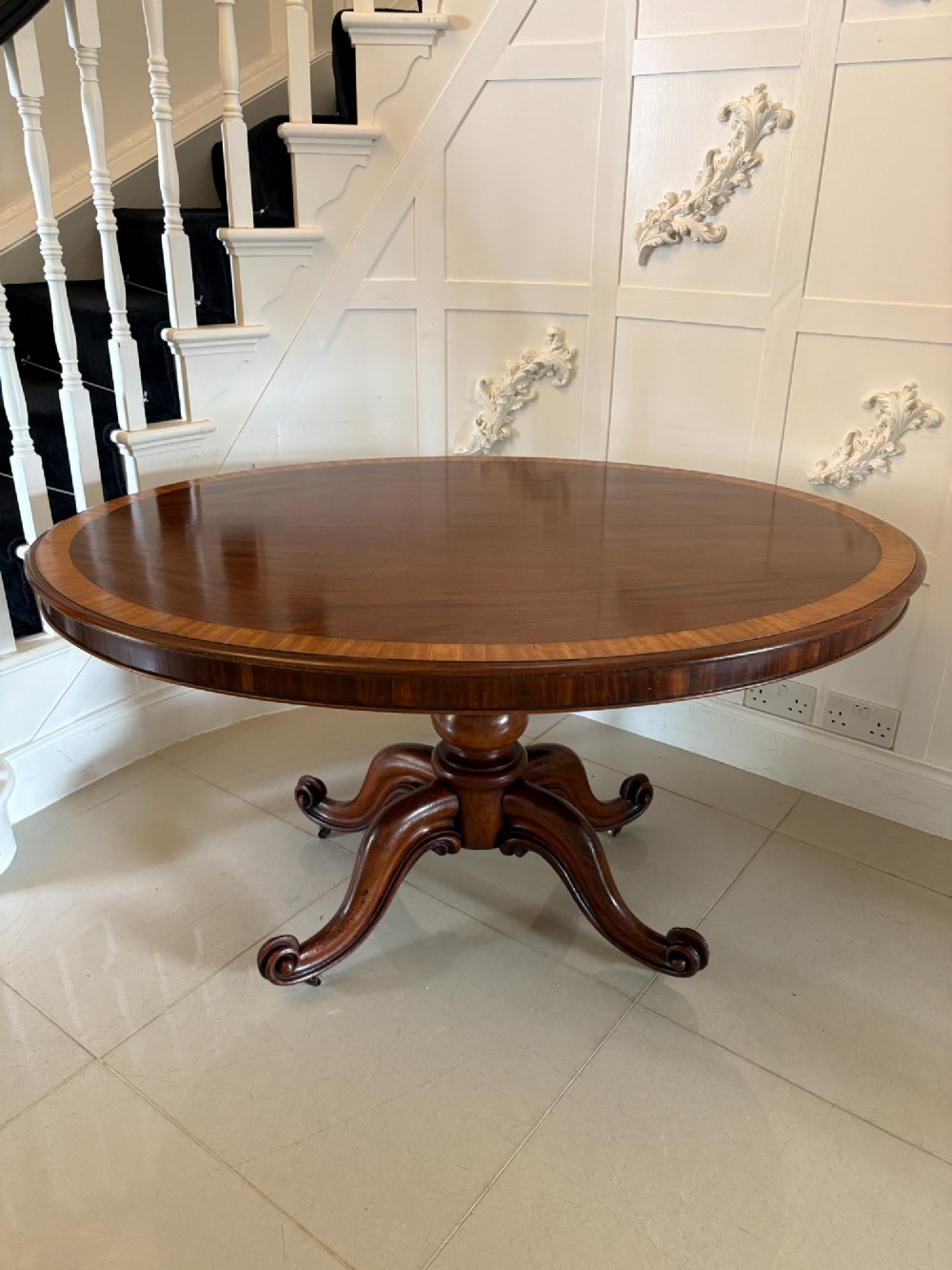 An oval Victorian tilt-top dining table in figured mahogany with satinwood crossbanding, set on a quadruped pedestal base with brass castors. View IV