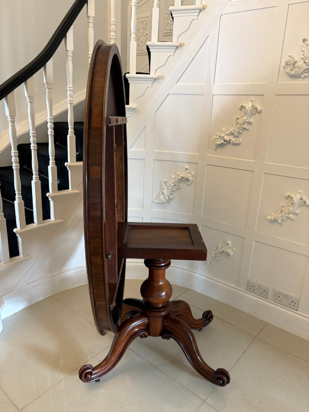 An oval Victorian tilt-top dining table in figured mahogany with satinwood crossbanding, set on a quadruped pedestal base with brass castors. View XI