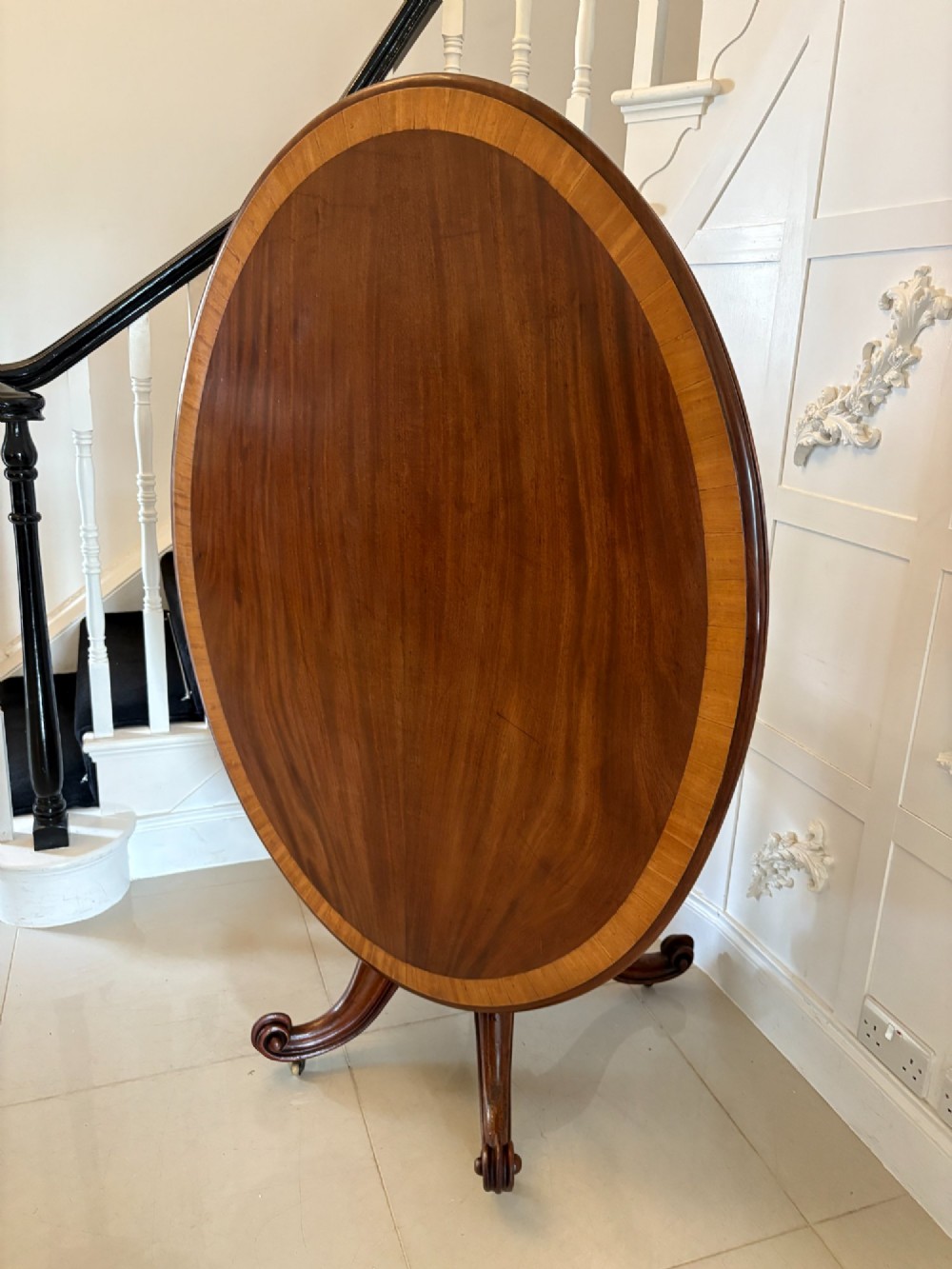 An oval Victorian tilt-top dining table in figured mahogany with satinwood crossbanding, set on a quadruped pedestal base with brass castors. View XII