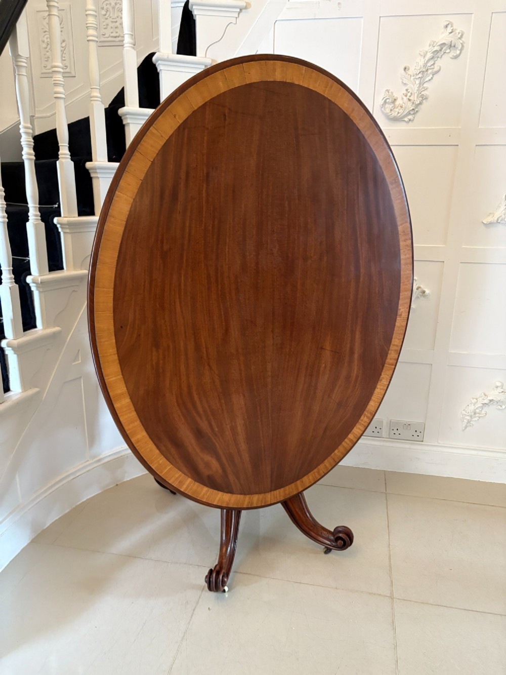 An oval Victorian tilt-top dining table in figured mahogany with satinwood crossbanding, set on a quadruped pedestal base with brass castors. View XIII