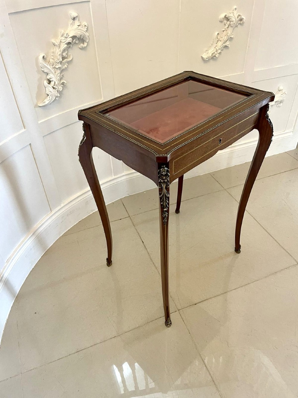 A 19th-century French mahogany bijouterie display table featuring a glazed top with brass inlay, a single front drawer, and slender cabriole legs with brass mounts. View X