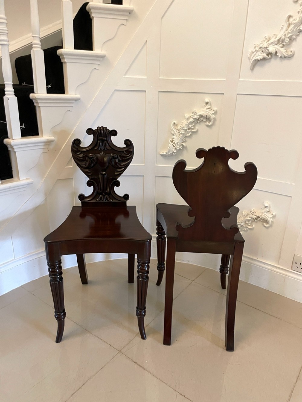 A pair of antique Regency dark mahogany hall chairs featuring scroll-carved cartouche backrests, solid timber seats, and reeded front legs. View XII