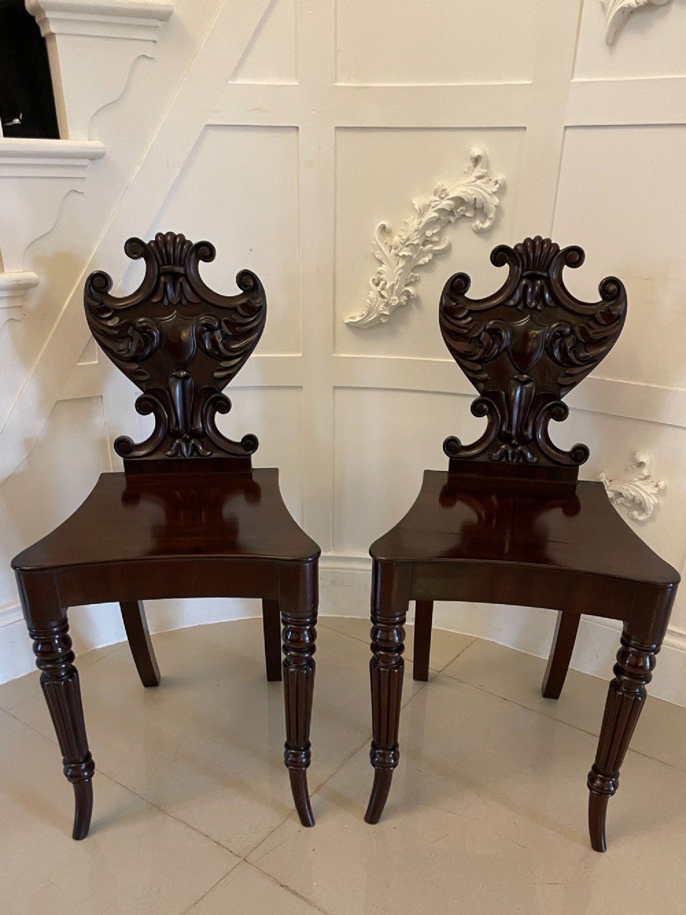 A pair of antique Regency dark mahogany hall chairs featuring scroll-carved cartouche backrests, solid timber seats, and reeded front legs.