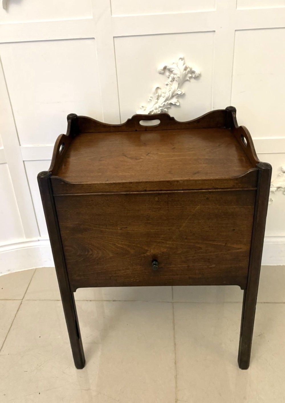 An 18th-century Georgian mahogany bedside cabinet featuring a gallery tray top with cut-out handles, a front cupboard door, and square legs.