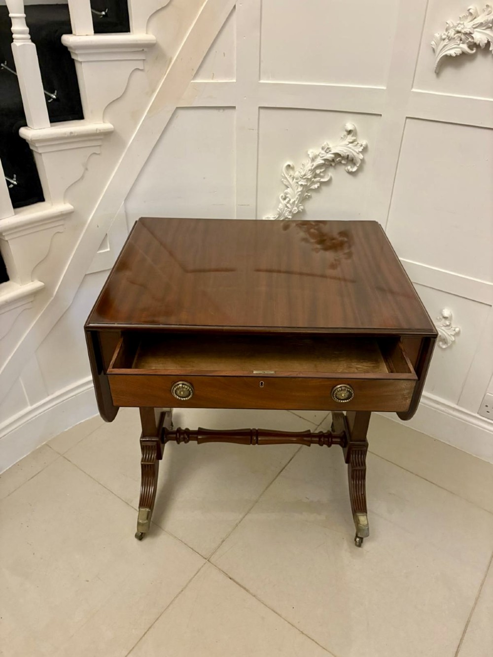 A Regency period mahogany sofa table featuring two drop-leaf sides, a single drawer with brass ring handles, reeded splayed legs, a turned stretcher bar, and brass castors. View II