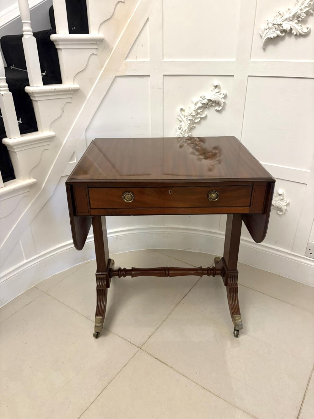 A Regency period mahogany sofa table featuring two drop-leaf sides, a single drawer with brass ring handles, reeded splayed legs, a turned stretcher bar, and brass castors.