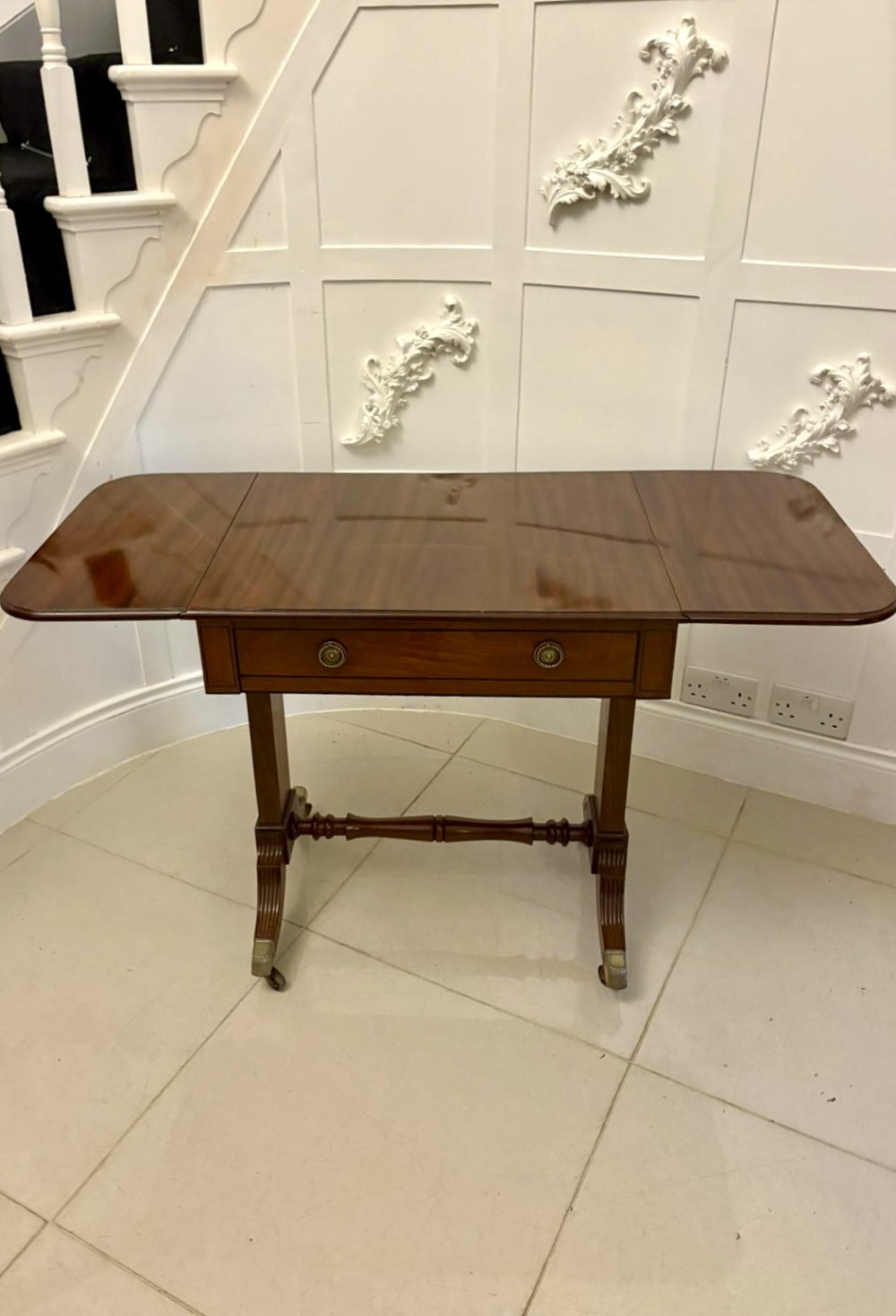 A Regency period mahogany sofa table featuring two drop-leaf sides, a single drawer with brass ring handles, reeded splayed legs, a turned stretcher bar, and brass castors. View VII