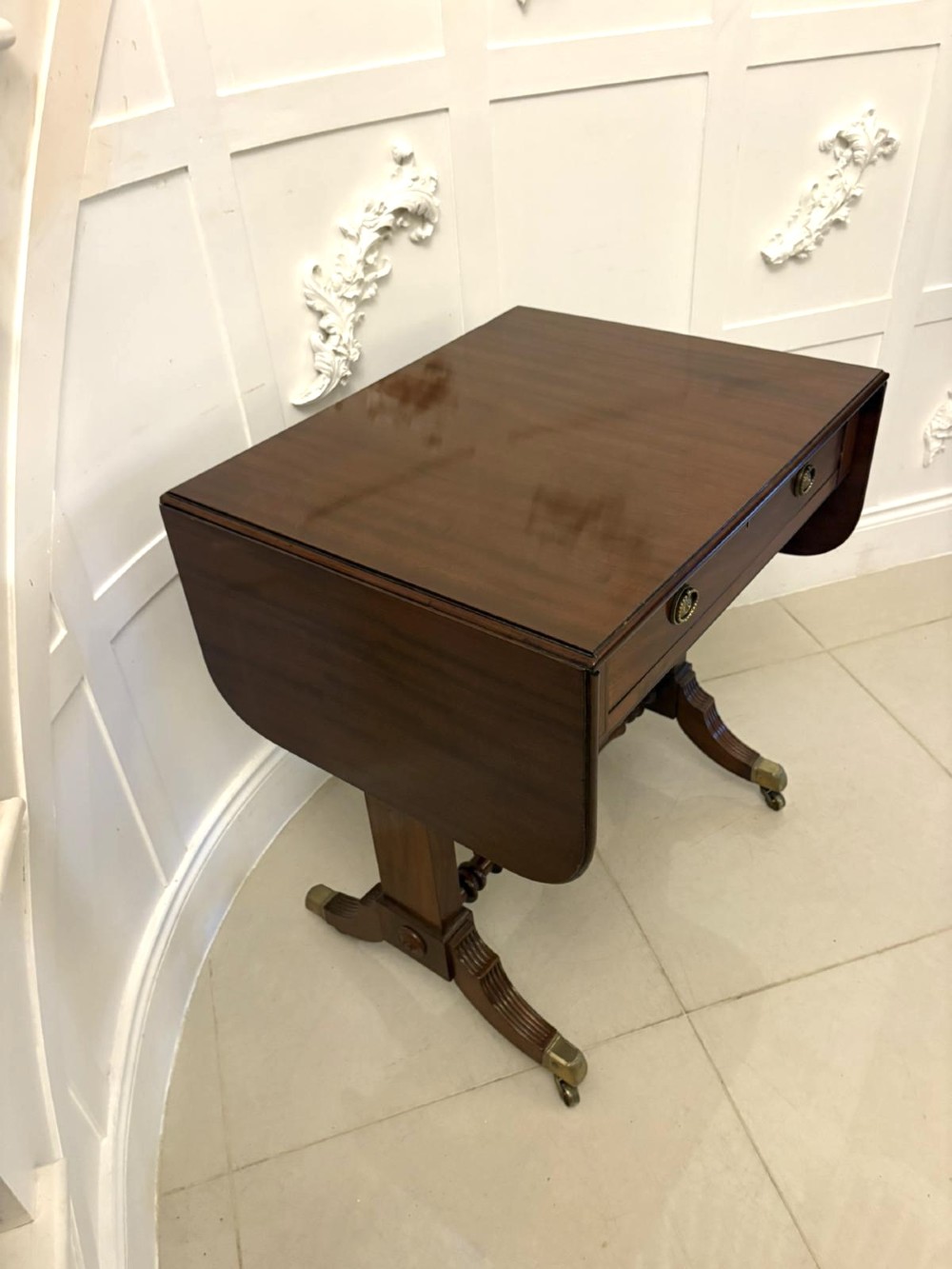 A Regency period mahogany sofa table featuring two drop-leaf sides, a single drawer with brass ring handles, reeded splayed legs, a turned stretcher bar, and brass castors. View VIII