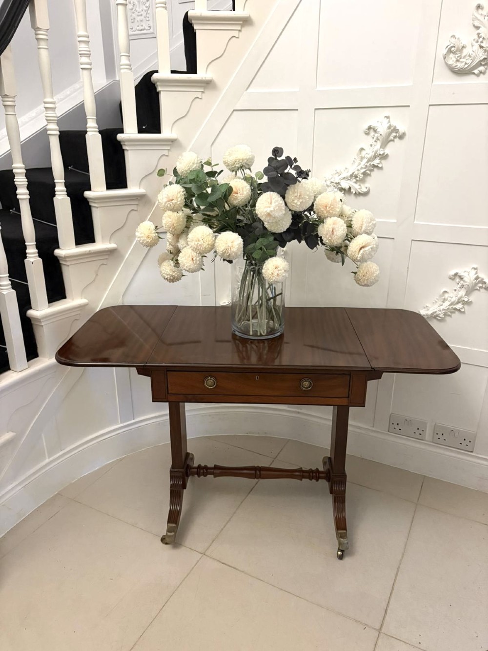 A Regency period mahogany sofa table featuring two drop-leaf sides, a single drawer with brass ring handles, reeded splayed legs, a turned stretcher bar, and brass castors. View X