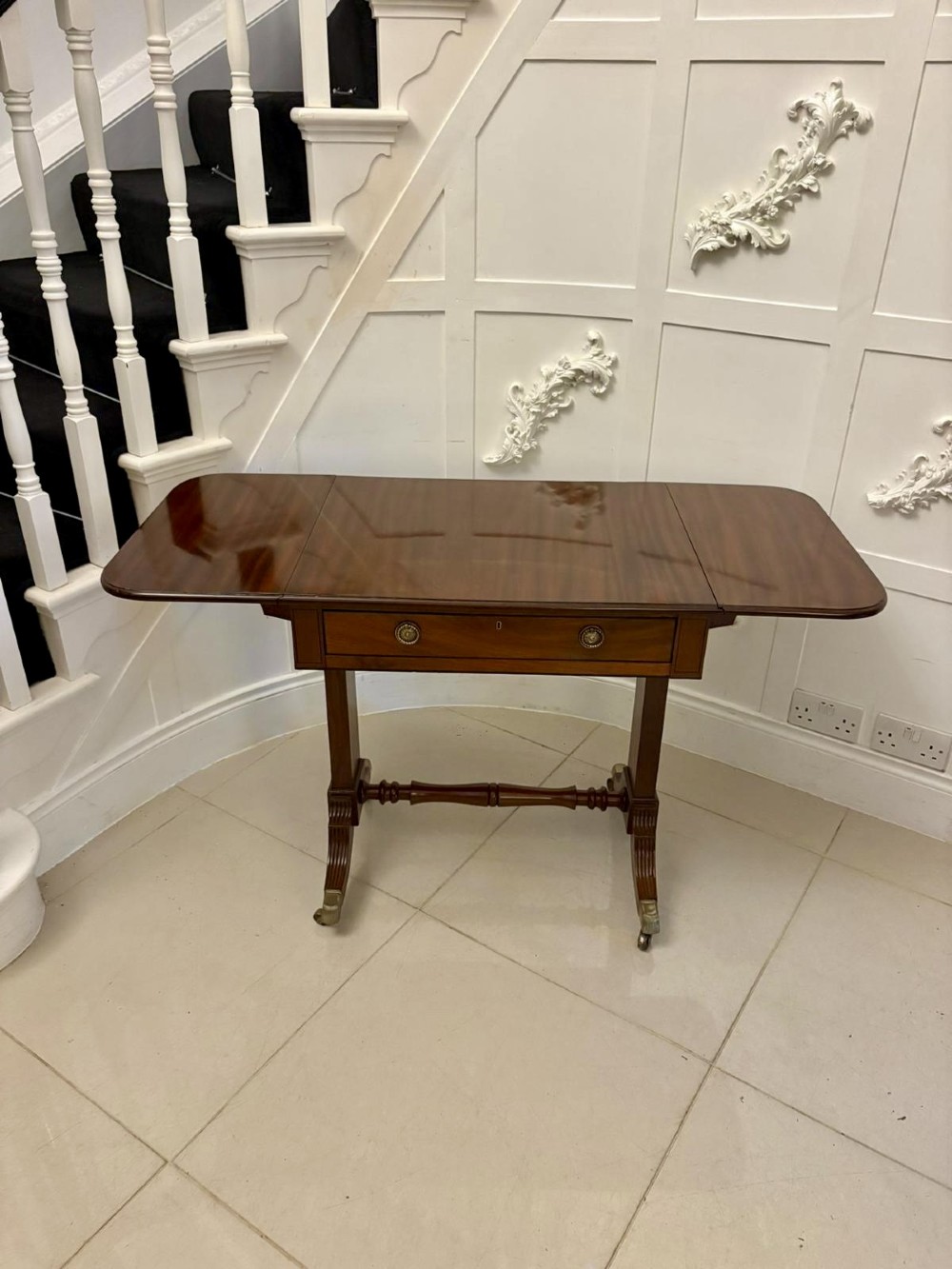 A Regency period mahogany sofa table featuring two drop-leaf sides, a single drawer with brass ring handles, reeded splayed legs, a turned stretcher bar, and brass castors. View XI