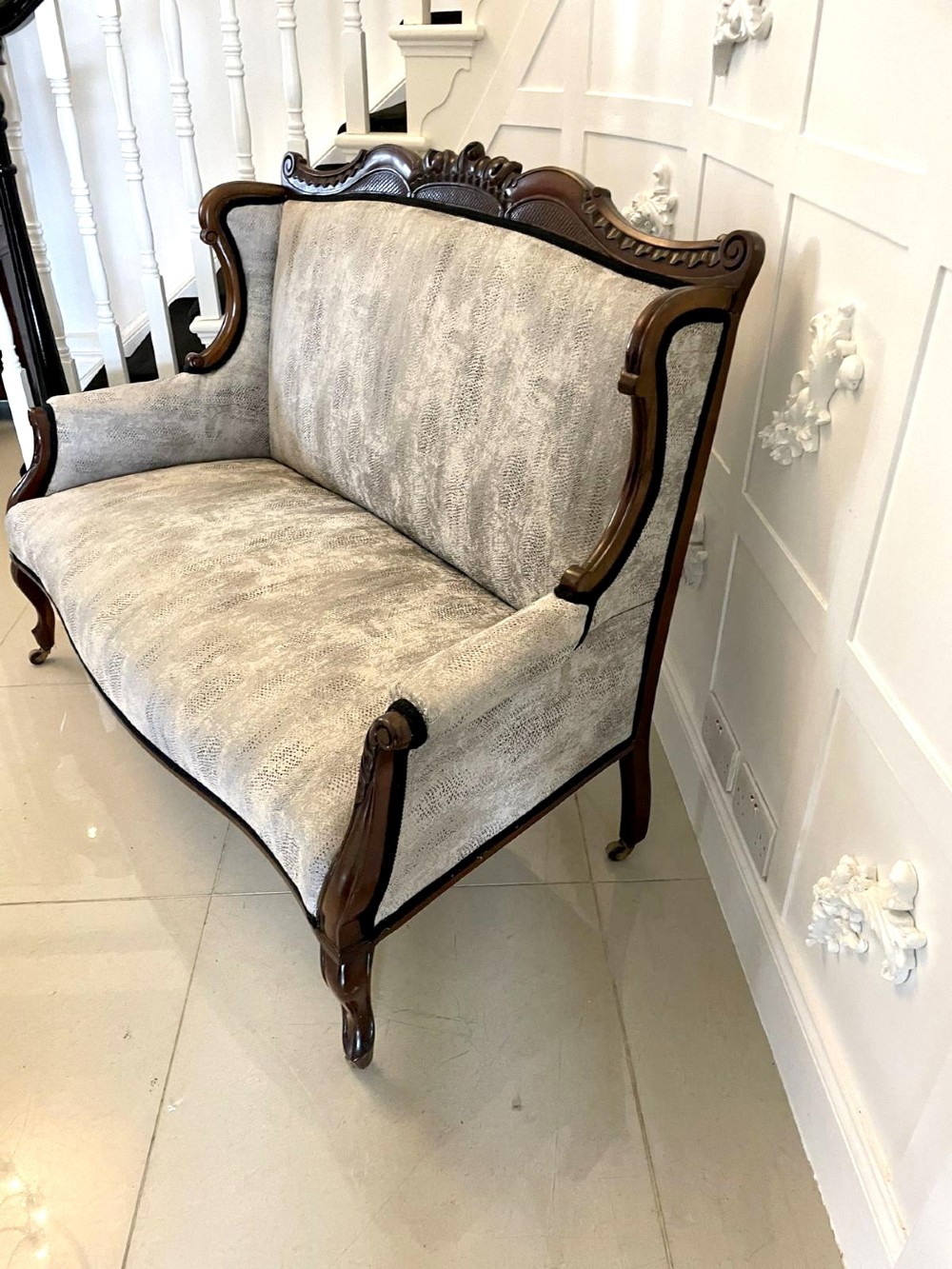 Victorian carved mahogany sofa featuring a decorative crest rail, curved scroll arms, grey textured upholstery, and cabriole legs on brass castors. View IV