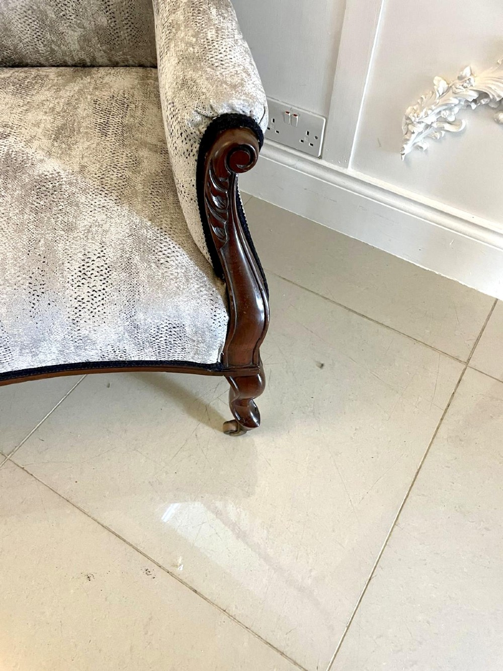 Victorian carved mahogany sofa featuring a decorative crest rail, curved scroll arms, grey textured upholstery, and cabriole legs on brass castors. View V
