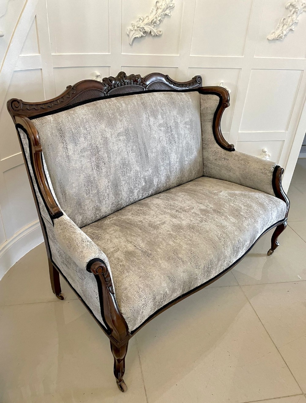 Victorian carved mahogany sofa featuring a decorative crest rail, curved scroll arms, grey textured upholstery, and cabriole legs on brass castors. View XIII