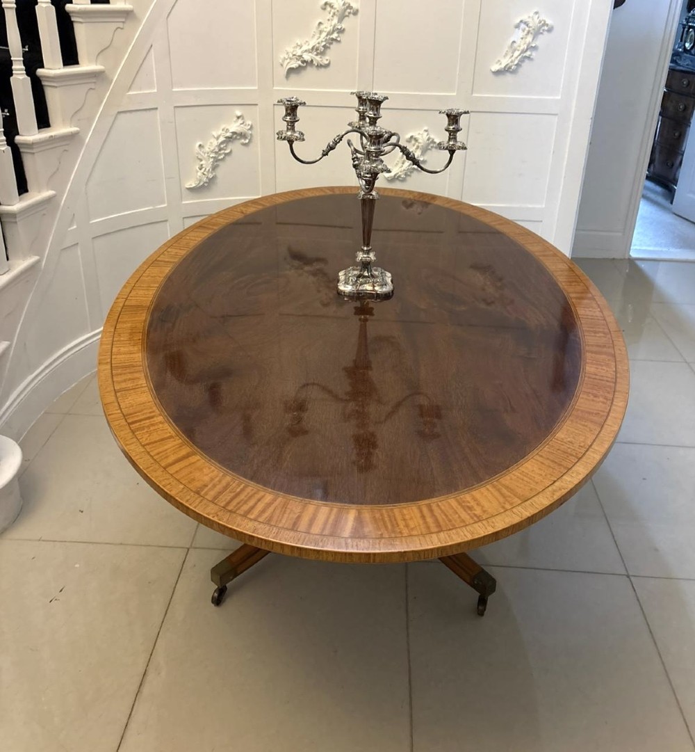 An antique Regency style oval mahogany tilt-top dining table featuring a figured timber top with satinwood crossbanding mounted on a pedestal base with brass castors. View X