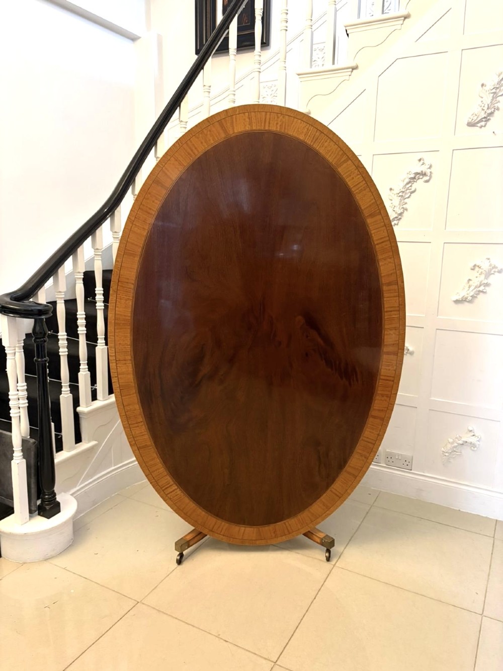 An antique Regency style oval mahogany tilt-top dining table featuring a figured timber top with satinwood crossbanding mounted on a pedestal base with brass castors.