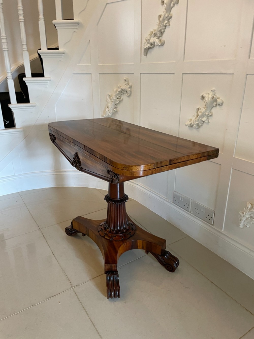An antique Victorian rosewood fold-over card table featuring a carved frieze, a fluted pedestal column, and a quadform base with scroll feet. View X