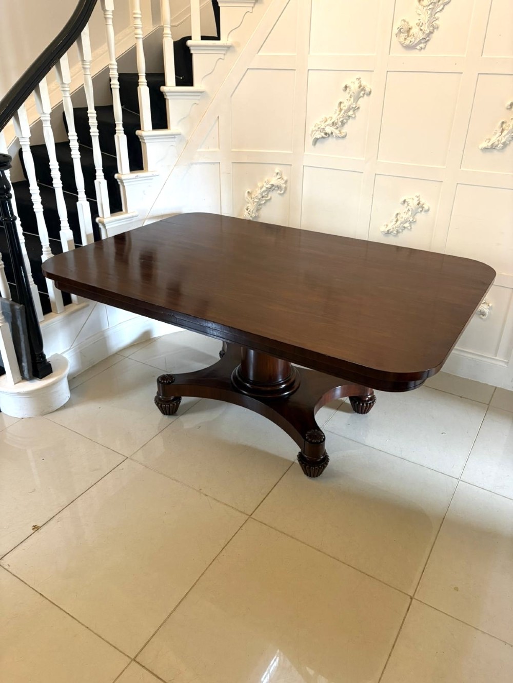 A William IV figured mahogany tilt top dining table shown upright, featuring rounded corners, a smooth grain finish, and a central pedestal base with carved feet. View VIII