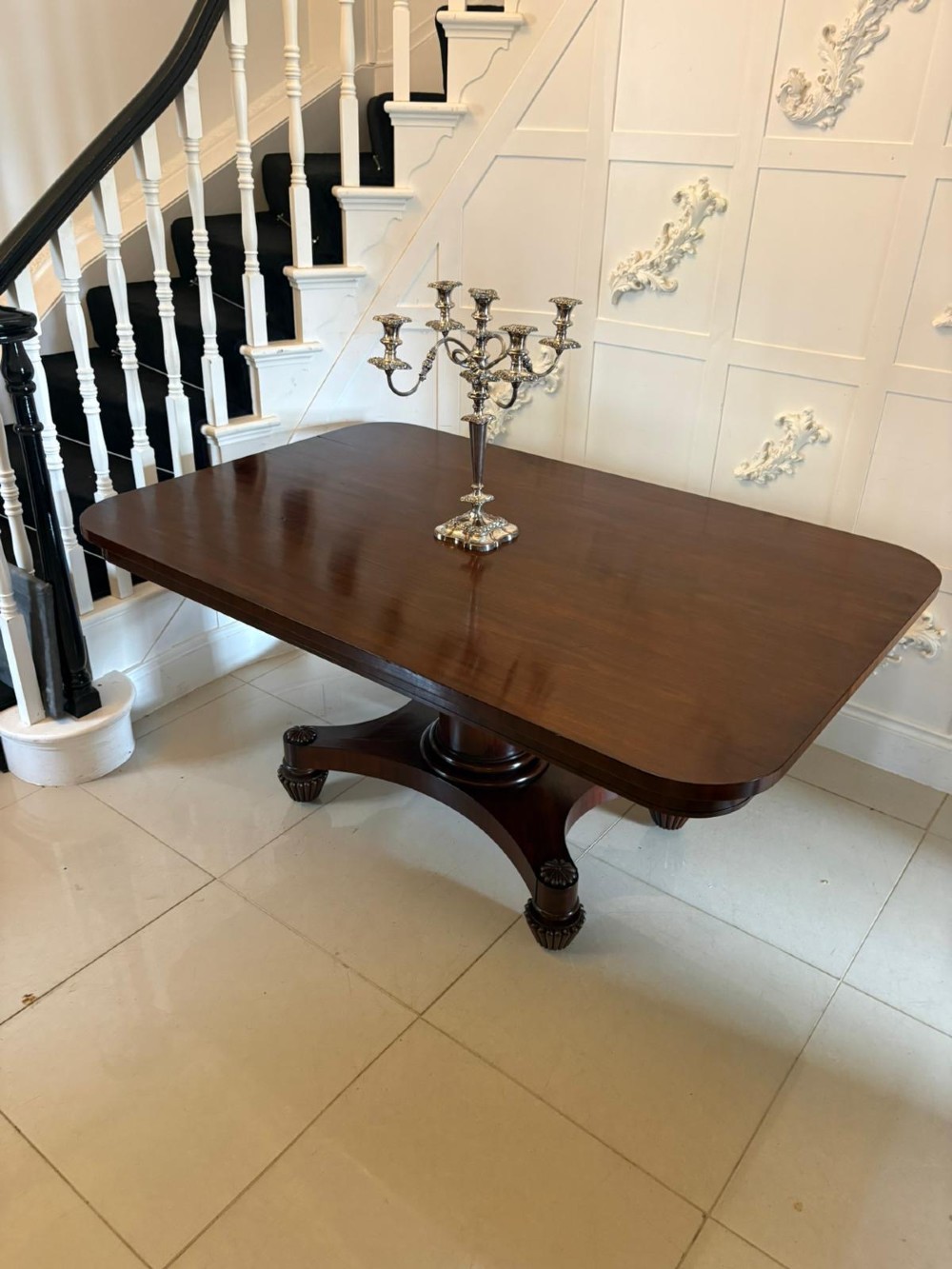A William IV figured mahogany tilt top dining table shown upright, featuring rounded corners, a smooth grain finish, and a central pedestal base with carved feet. View IX