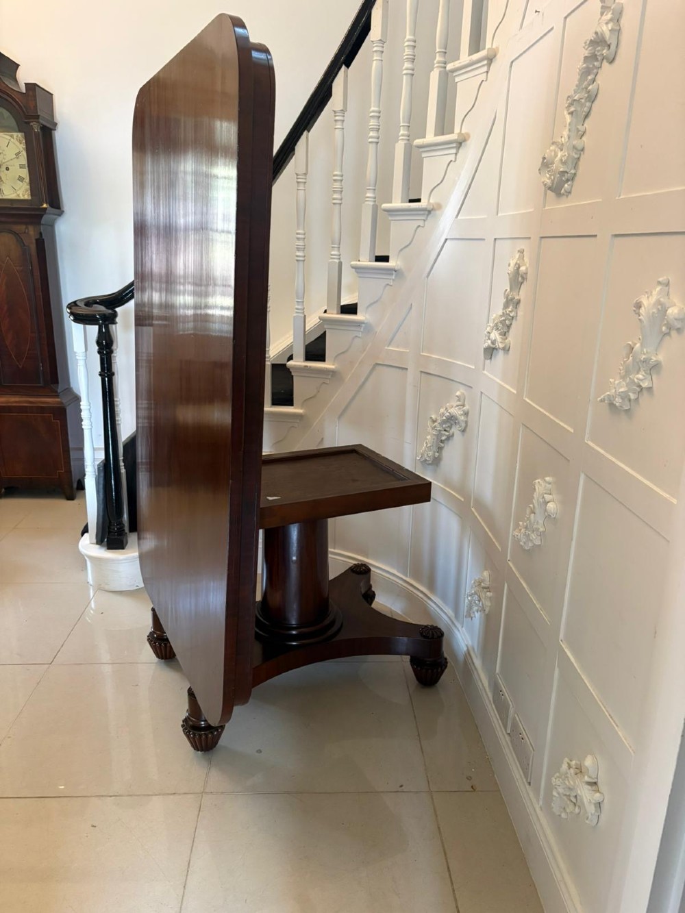 A William IV figured mahogany tilt top dining table shown upright, featuring rounded corners, a smooth grain finish, and a central pedestal base with carved feet. View X