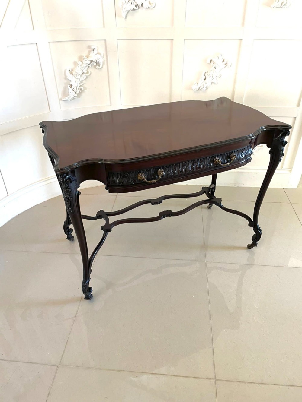 An antique Victorian dark mahogany centre table featuring a shaped serpentine top, carved frieze with brass handles, cabriole legs, and an arched scroll stretcher.