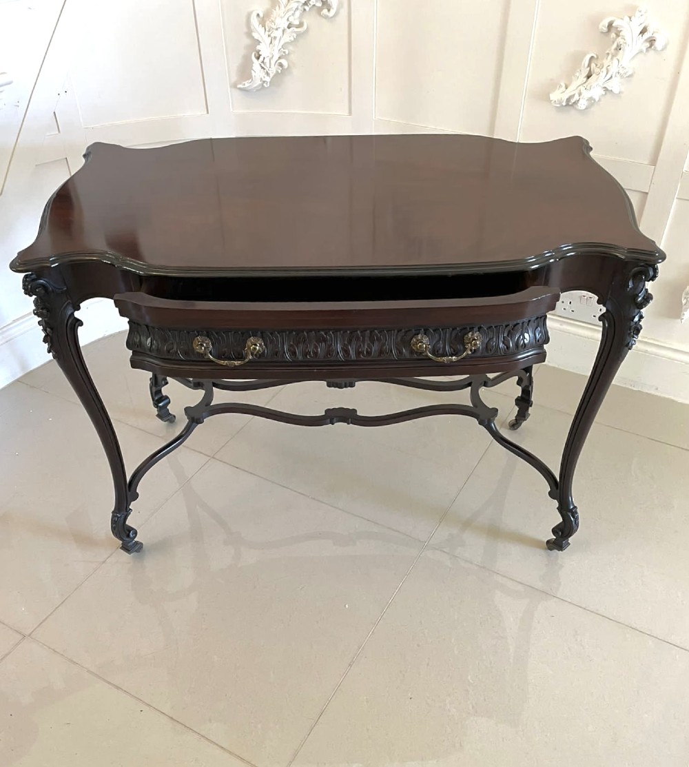An antique Victorian dark mahogany centre table featuring a shaped serpentine top, carved frieze with brass handles, cabriole legs, and an arched scroll stretcher. View X