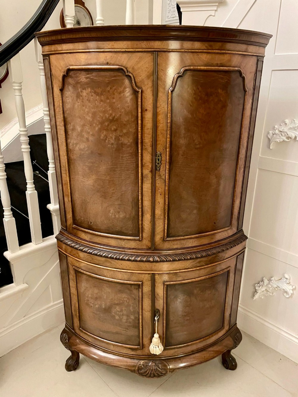An antique burr walnut bow-fronted cocktail cabinet featuring two upper doors, two lower doors, carved cabriole legs, and brass keyhole escutcheons.