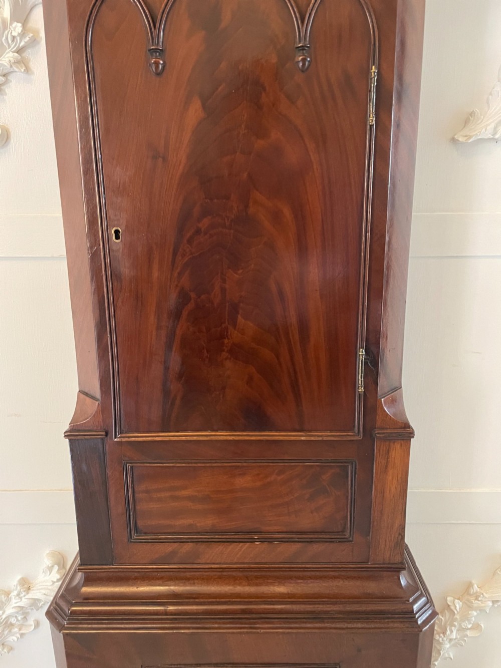 A George III flame mahogany longcase clock featuring a swan-neck pediment, turned hood columns, a painted arch dial, and a gothic-arched trunk door. View V