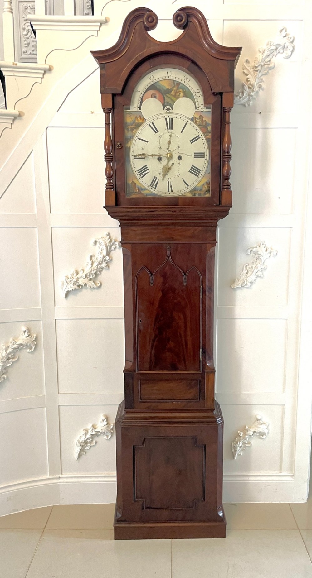 A George III flame mahogany longcase clock featuring a swan-neck pediment, turned hood columns, a painted arch dial, and a gothic-arched trunk door.