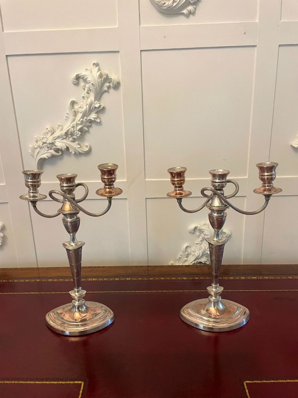 A pair of antique silver-plated three-light candelabras with curved branch arms, tapered baluster stems, and moulded oval bases showing subtle copper undertones.